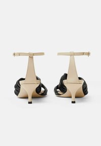 Czarne i beżowe sandały na wysokim obcasie z paskiem na kostkę, z falbanką z czarnej skóry i smukłym, kontrastującym obcasem.