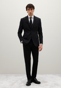 Completo nero su misura con giacca aderente, rever a punta e due bottoni, abbinato a pantaloni coordinati e una camicia bianca. Scarpe di pelle.