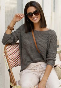 Grauer gestrickter Pullover mit lockerer Passform, kombiniert mit hellbeigen Hosen. Übergroße schwarze Sonnenbrille und eine braune Umhängetasche vervollständigen das Outfit.