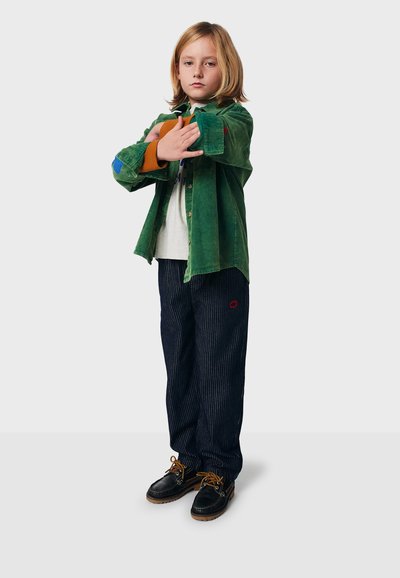 Kind mit blonden Haaren, das eine grüne Jacke trägt, die Ärmel hochkrempelt, ein weißes Hemd und gestreifte dunkle Hosen sowie schwarze Schuhe trägt, steht vor einem einfarbigen Hintergrund.