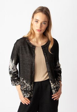 Cardigan noir avec imprimé floral blanc sur les manches, col rond et devant à boutons. Texture douce, avec manches longues et coupe décontractée.