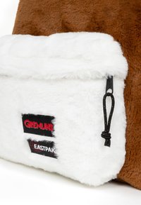 Borsa in materiale di pelliccia marrone con una tasca frontale bianca e soffice. Presenta i loghi "Gremlins" e "Eastpak" su etichette in tessuto nero. Corda nera di estrazione attaccata.