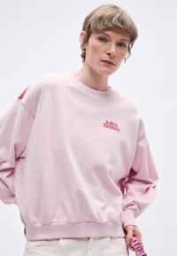 Ružová mikina s rebrovaným okrúhlym výstrihom a elastickým lemom. Na hrudi je malý červený logo a na ramene kontrastná červená záplata.