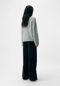 Helle graue, übergroße Strickjacke mit einer weichen Textur, kombiniert mit hoch taillierten, schwarzen weit geschnittenen Hosen, von hinten vor einem weißen Hintergrund sichtbar.