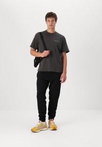 Camiseta gris con el logotipo "Dickies", pantalones de chándal negros y zapatillas amarillas/ grises. Bandolera negra llevada en cruz. Atuendo casual con elementos deportivos.