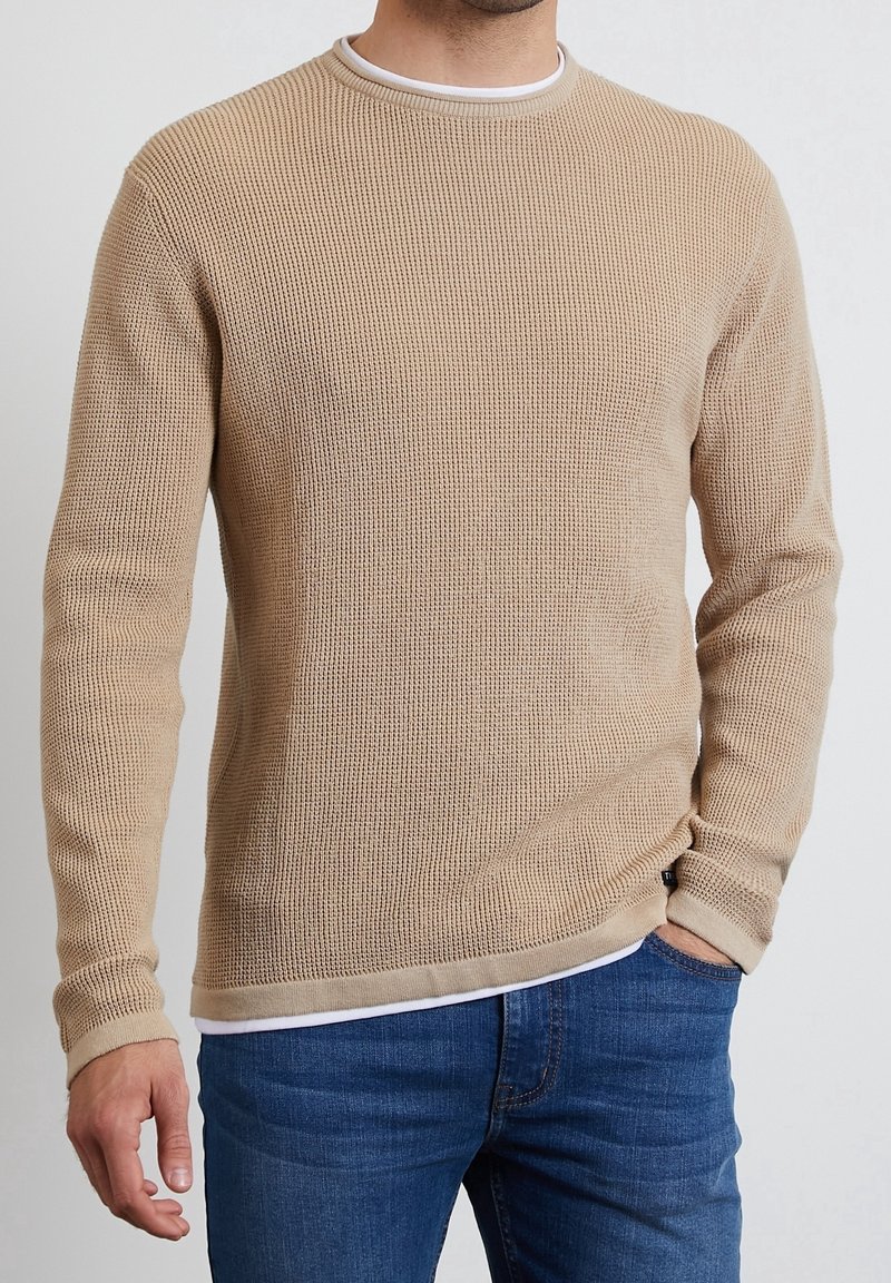Pull en laine beige avec des manches longues, un col rond, une texture côtelée, et un col intérieur blanc contrastant. Porté sur un jean bleu.