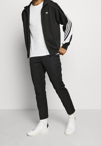 Zwarte zip-up jack met witte streepaccenten, gecombineerd met zwarte broeken en witte sneakers. De outfit heeft een casual sportief ontwerp.