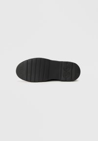 Semelle de chaussure en caoutchouc noir présentant des rainures horizontales et des surfaces texturées, avec un logo sur le côté pour une meilleure adhérence et durabilité.
