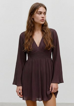 Femme aux longs cheveux bruns ondulés portant une robe plissée foncée à manches longues avec un décolleté en V, debout devant un fond clair uni.