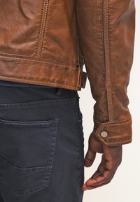 Veste en cuir marron avec une finition texturée, présentant des surpiqûres contrastées et un détail de bouton-pression au niveau du poignet. Portée sur un pantalon en tissu foncé.