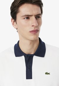 Polo con cuello y botones azul marino, cuerpo blanco, tejido texturizado y un pequeño logotipo de cocodrilo verde en el lado izquierdo del pecho.