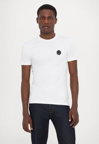 Hvid kortærmet T-shirt lavet af bomuld, med rund halsudskæring og et cirkulært sort logo på venstre bryst. Sæt sammen med mørkeblå jeans.