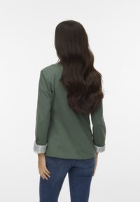 Blazer verde con maniche a righe arrotolate, design aderente e texture liscia. Indossato sopra jeans blu, visto da dietro.