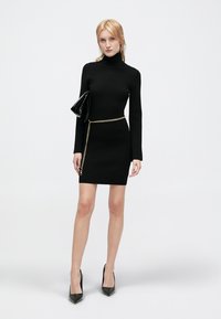Fekete bordázott kötött ruha magas nyakkal, hosszú ujjakkal és formázott siluettel. Arany láncövvel és fekete táskával kiegészítve.