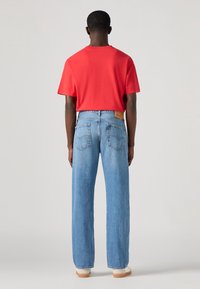 Uomo in piedi di spalle, che indossa una maglietta rossa, jeans dritti azzurri e scarpe bianche, su uno sfondo neutro.