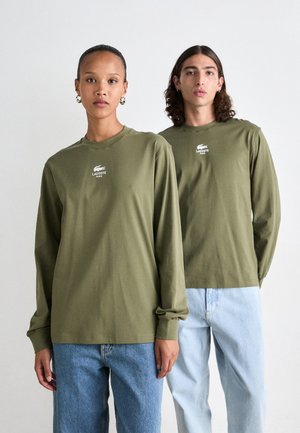 Deux modèles sont debout, portant des chemises à manches longues Lacoste vert olive assorties avec des jeans bleu clair, devant un fond blanc uni.