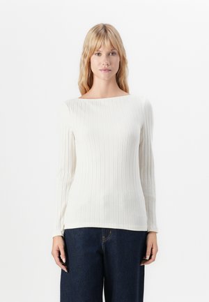 Donna con capelli biondi che indossa un top lungo a maniche ribattuto color crema e jeans blu scuro, in piedi contro uno sfondo bianco semplice.