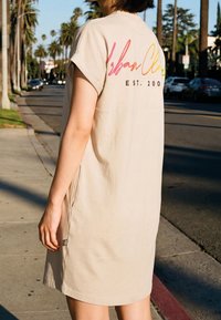 Personne portant une robe ample beige avec un texte multicolore dans le dos, debout sur un trottoir ensoleillé avec des palmiers et des voitures garées à proximité.