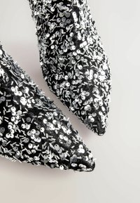 Mustad mustreeritud mustad kõrge kontsaga kingad, millel on hõbedased sära ja metalldetailid, ning terava nina disain.