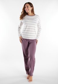 Woman smiling, standing barefoot wearing white long-sleeve striped top and mauve full-length pants against a white background.