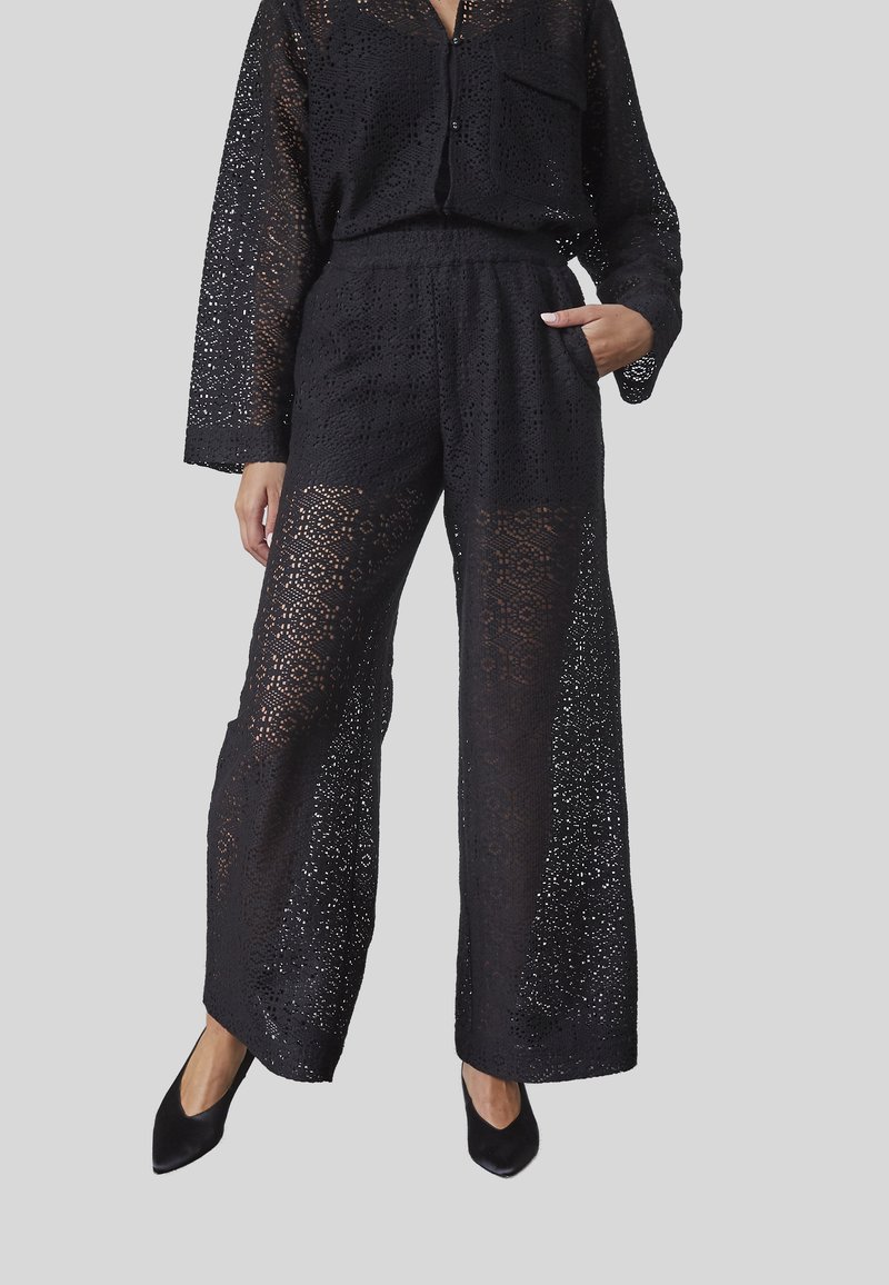 Sæti i sort guipure blonder bestående af en langærmet skjorte med knaplukning og vide bukser, der fremhæver indviklede mønstre og en gennemsigtig tekstur.