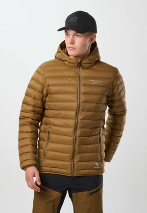 Homme portant une veste matelassée marron avec capuche et casquette noire, légèrement tourné vers la gauche avec les mains près des côtés, sur fond gris clair.