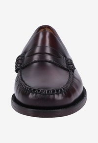 Mocasín de cuero marrón con un acabado suave, detalles decorativos de costura y punta redondeada. Presenta una suela baja y plana de color negro.