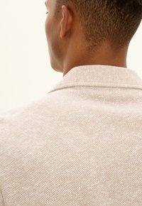 Beige texturerad kavaj med krage, slät tyg och synliga sömmar. Bakifrån framhävs halslinjen och det subtila tygmönstret.
