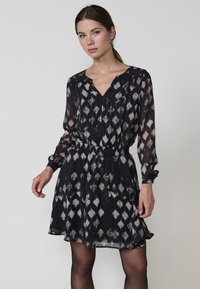 Schwarzes und weißes gemustertes Kleid aus leichtem Stoff, mit V-Ausschnitt, langen Ärmeln und elastischem Taillenbund.