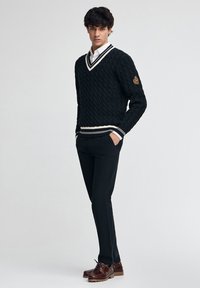 Svart stickad tröja med kabelmönster, V-ringning och vita randiga manschetter; matchad med svarta byxor och bruna skor.