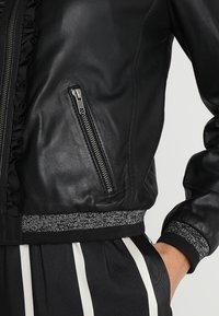 Czarna skórzana kurtka bomber z marszczonymi detalami, zapinana na zamek kieszeń boczna oraz teksturowany szary dzianinowy dół i mankiety.