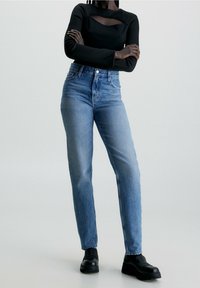 Niebieskie dżinsy z wysokim stanem o prostych nogawkach, z delikatnym wyblaknięciem, pięciokieszeniowym fasonem i zapięciem na guziki. Czarnie botki do kostki uzupełniają stylizację.