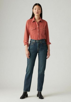 Camisa de manga larga color óxido con dos bolsillos en el pecho, combinada con vaqueros de tiro alto en azul oscuro y botas negras tipo botín.