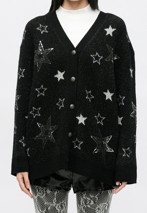 Cardigan noir boutonné avec des motifs d’étoiles blanches et noires, porté sur un top blanc et associé à des leggings à motif dentelle.