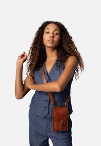 Brun läder crossbody-väska med spänne, med böjda kanter och sömnadsdetaljer, bärs av en modell i en blå outfit.