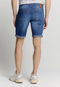 Denim shorts in medium blauw, met een opgerolde zoom, achterzakken en een leren label op de tailleband. Draagt een lichte grijze sportschoenen.