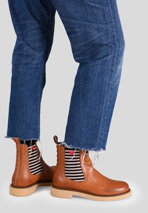 Stivaletti in pelle marrone con pannelli elastici laterali, suola beige chiaro e cuciture rosse. Jeans blu con orlo sfilacciato e calzini a righe bianche e nere.