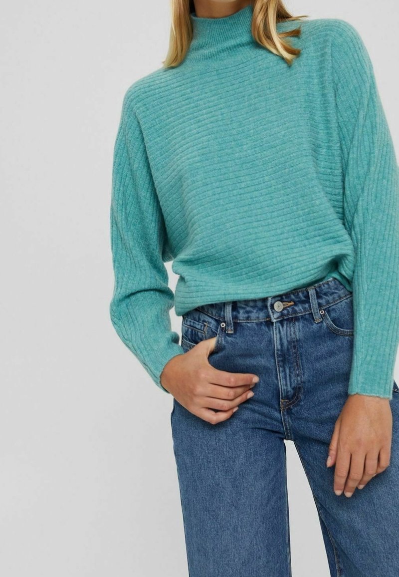 Pull côtelé turquoise clair avec col montant et manches longues, associé à un jean bleu taille haute. La texture semble douce.