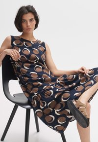 Navy dress with brown and cream circular patterns; sleeveless design, relaxed fit, gathered waist detail, paired with brown footwear.