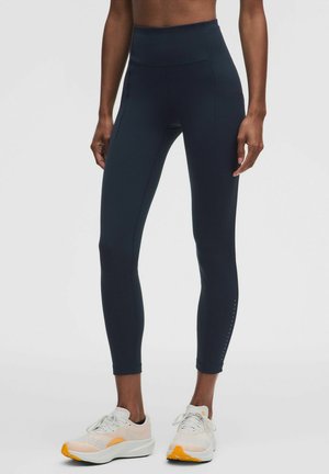 Person trägt schwarze Leggings mit hoher Taille und weiße Sportschuhe mit orangen Sohlen, steht vor einem einfarbigen hellen Hintergrund.