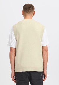 Una persona è in piedi con la schiena rivolta verso la camera, indossando un maglione smanicato beige sopra una camicia bianca e pantaloni scuri.