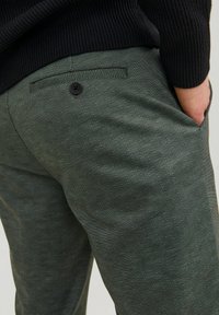 Pantaloni a motivi a spina di pesce verdi con una tasca posteriore, chiusura con bottone nero e tessuto texturizzato. Mano parzialmente visibile in tasca.