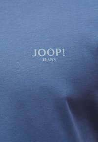 Ανοιχτό μπλε ύφασμα με την επιγραφή "JOOP! JEANS" σε λευκό, κεντραρισμένη στην επιφάνεια, εμφανίζοντας την υφή του υφάσματος και ελαφριές πτυχώσεις.