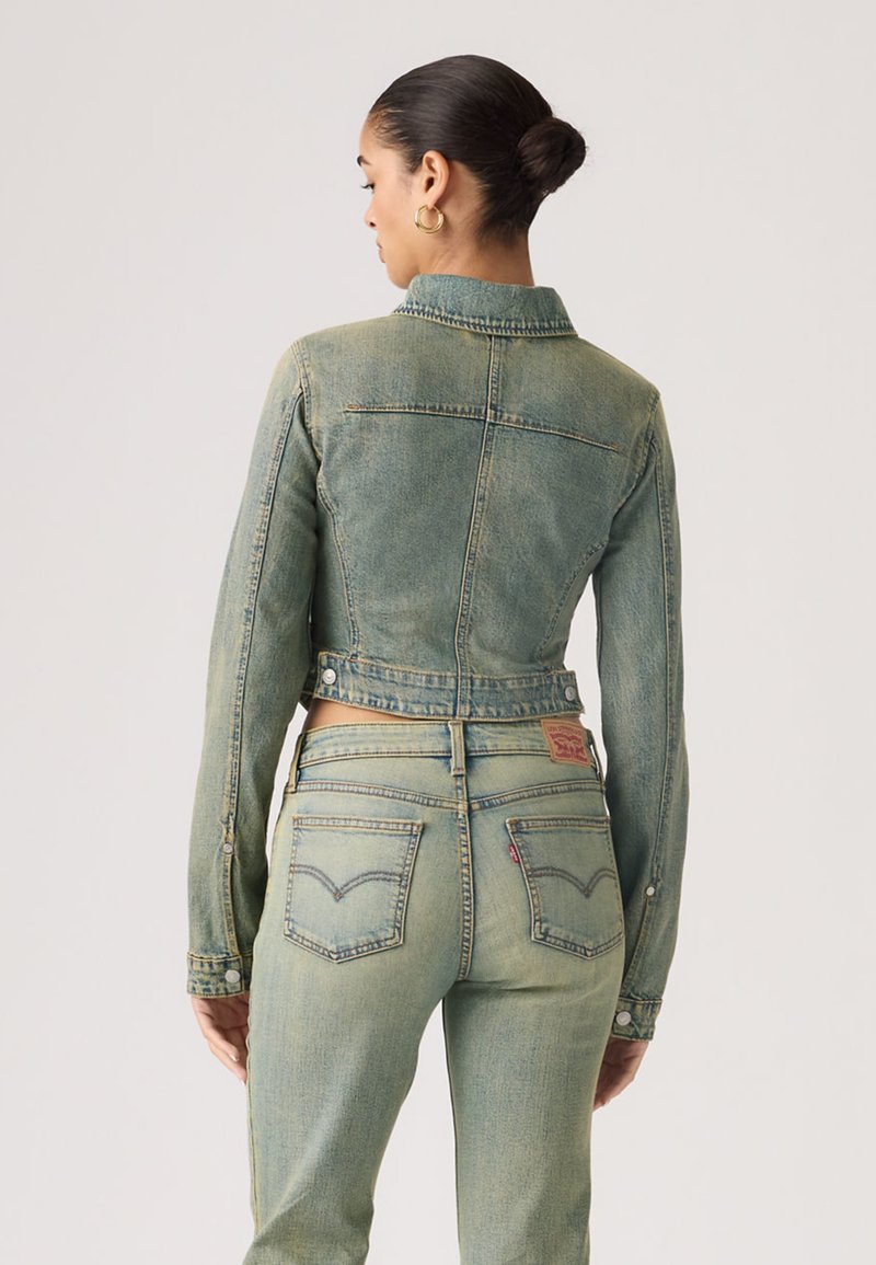 Femme avec les cheveux en chignon portant des boucles d'oreilles créoles dorées, une veste en jean délavée et un jean taille haute assorti, vue de dos sur un fond uni.