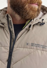 Chaqueta de plumas beige con costuras diagonales, cremallera negra y texto gris llamativo. Cuenta con una capucha con forro amarillo neón.