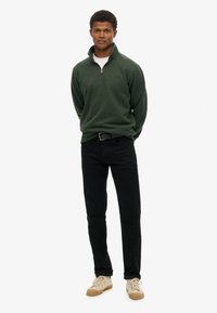 Donkergroene fleece trui met een kwart rits, gedragen over een wit shirt, gecombineerd met zwarte jeans en beige veterschoenen.