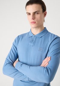 Dlhý rukáv polos tričko v svetlom modrom prevedení. Obsahuje golier, dve gombíky a embosované logo na hrudi. Mäkka a priedušná látka.