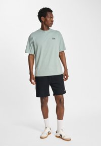 T-shirt vert clair à manches courtes avec un petit logo, associé à un short noir. Le modèle porte des baskets blanches avec des rayures noires et des chaussettes blanches.