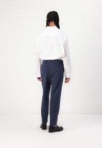 Camisa blanca de manga larga con botones y con un yugo visible, combinada con pantalones de vestir azul marino que tienen dos bolsillos traseros, llevada con zapatos negros.