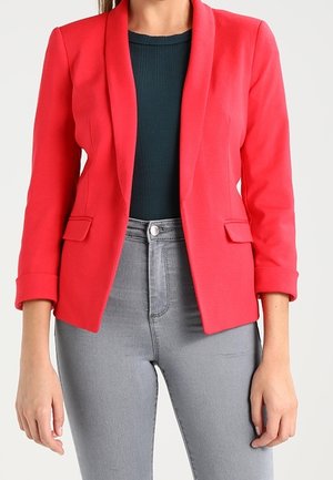 Kobieta ubrana w dopasowaną czerwoną marynarkę z podwiniętymi mankietami, na ciemnym, prążkowanym topie i jasnoszarych, wysokowysokościowych dżinsach.